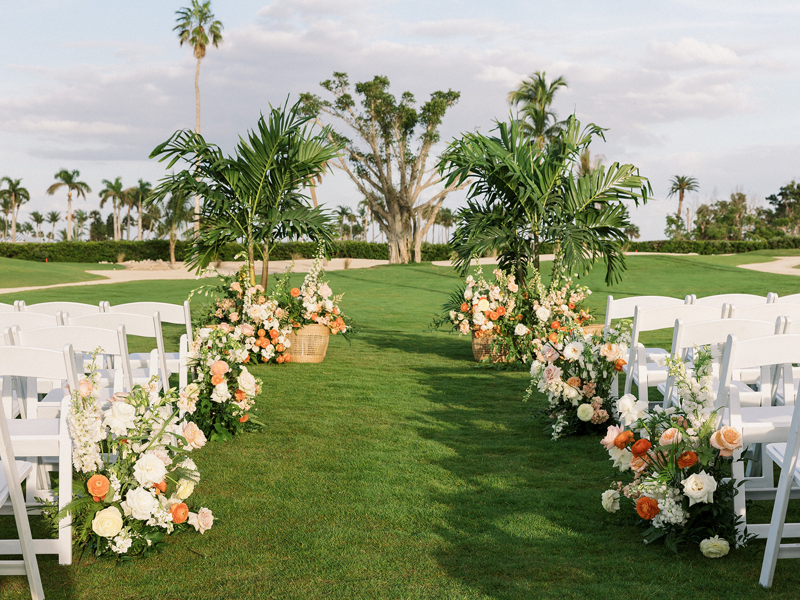 Wooden white chairs decorated with flowers on grass lawn with palm trees ready for a wedding ceremony.