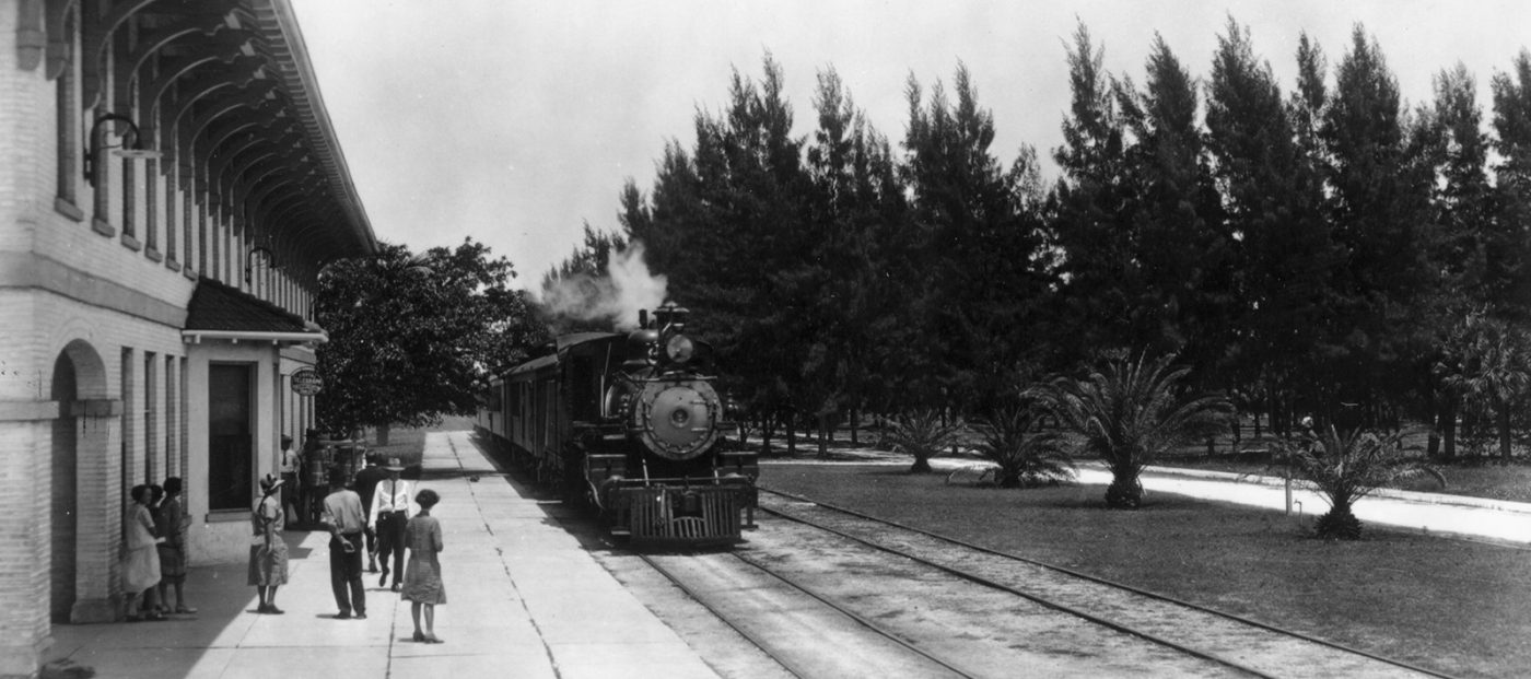A steam train arrives at a station where a small group is waiting