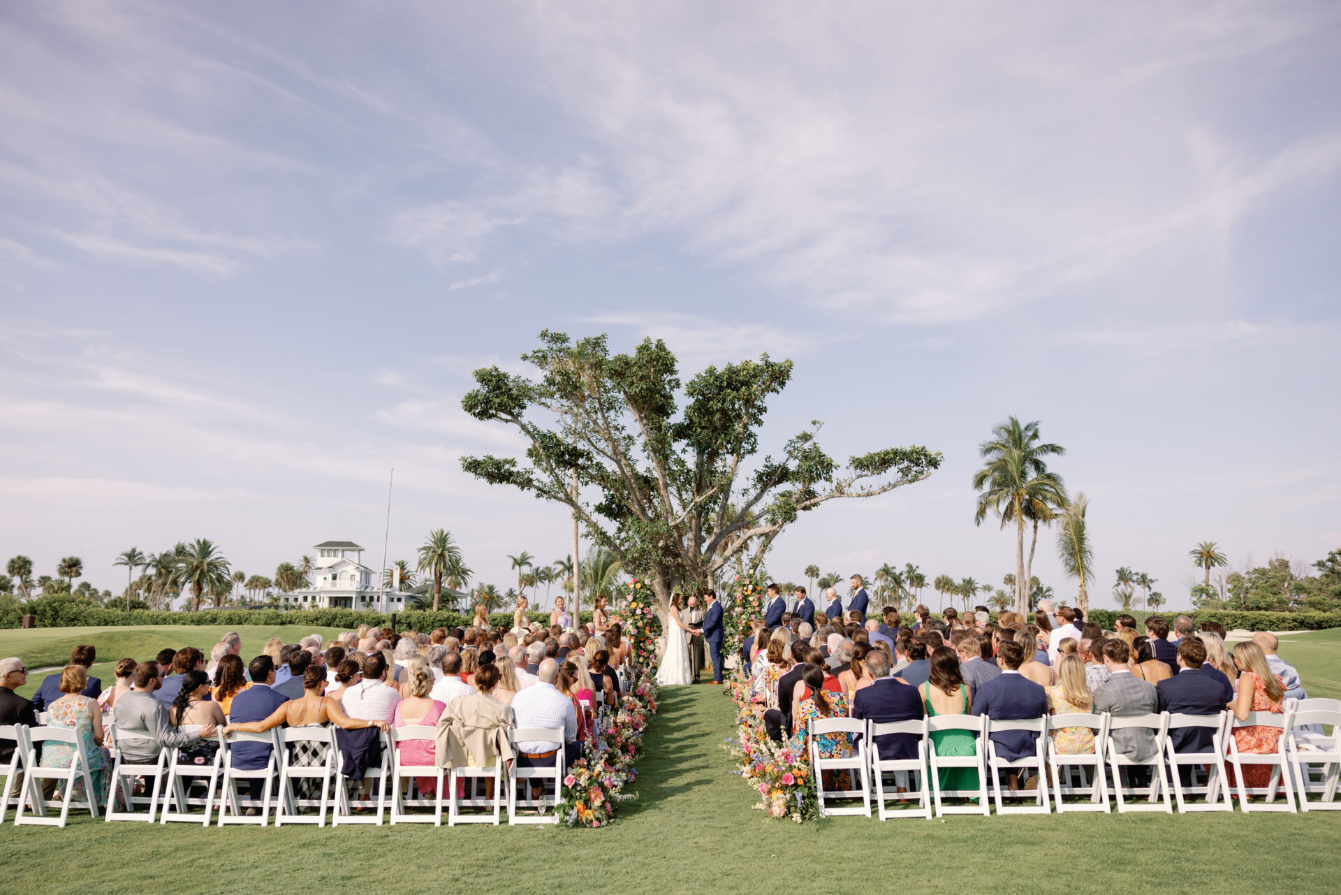 Couple takes their vows on grass lawn with a crowd watching from white chairs.