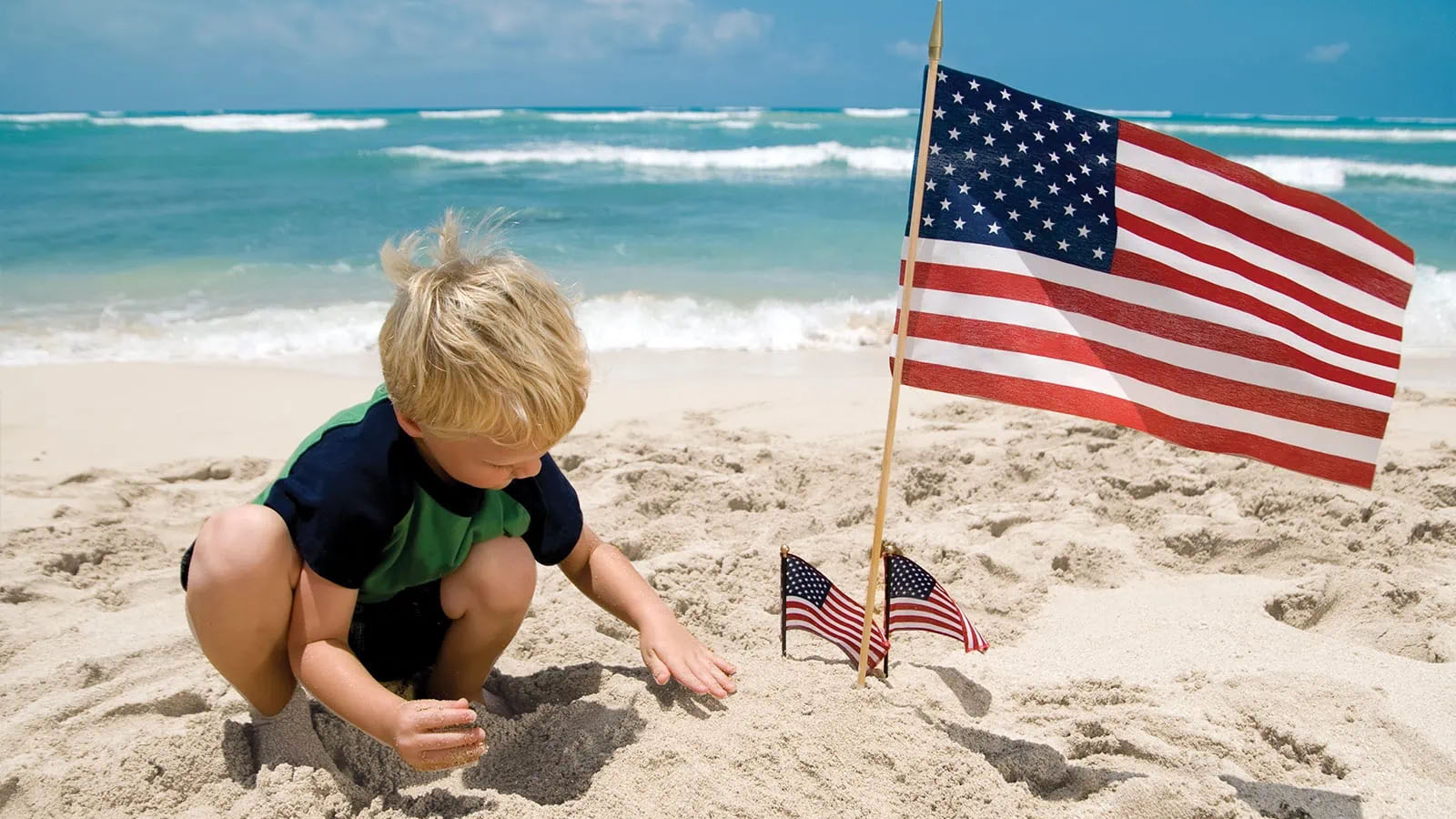 Little boy builds sandcastle on beach amidst small American flags