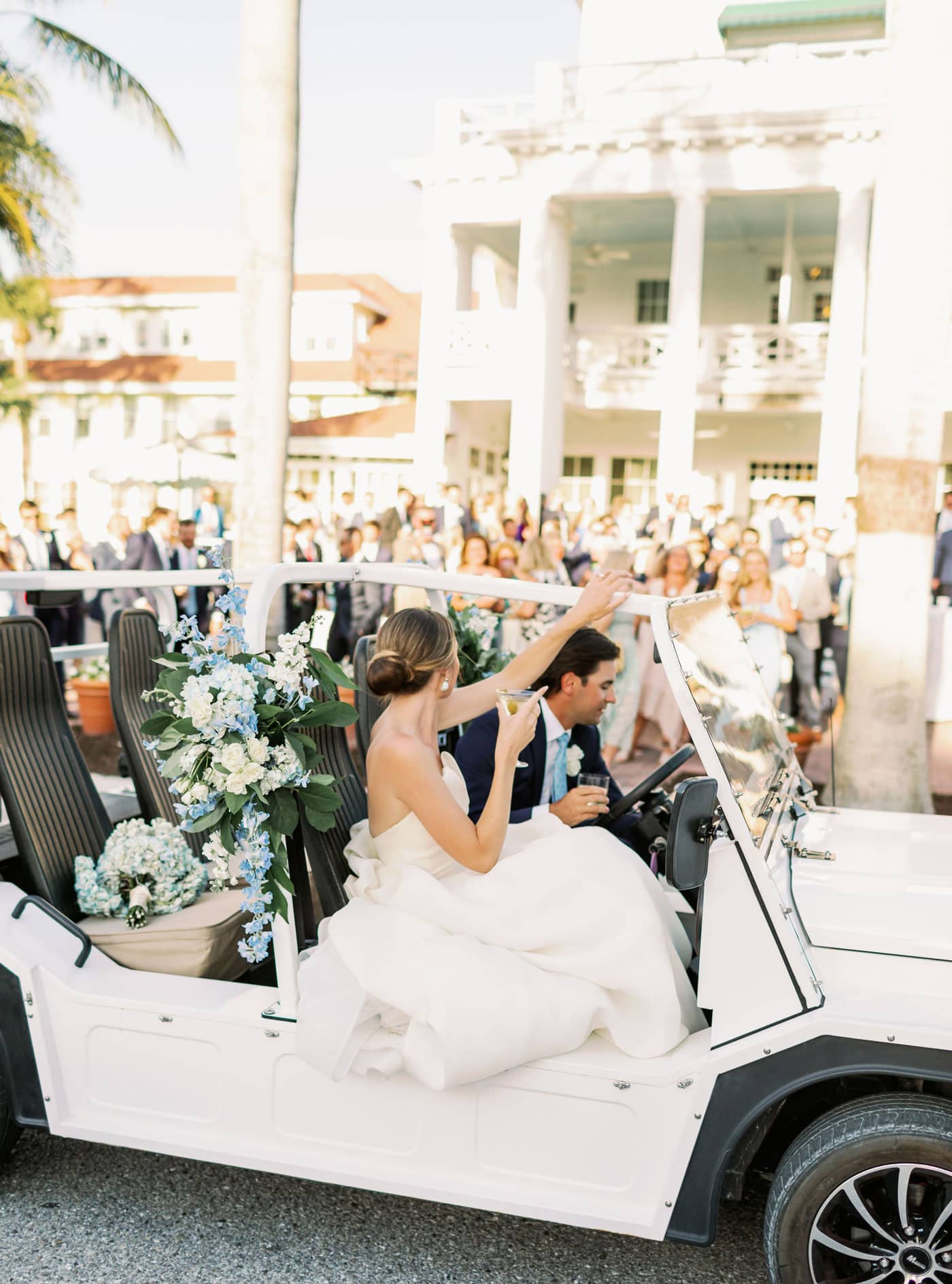 Bride and groom in a decorated golf cart wave goodbye to a cheering crowd