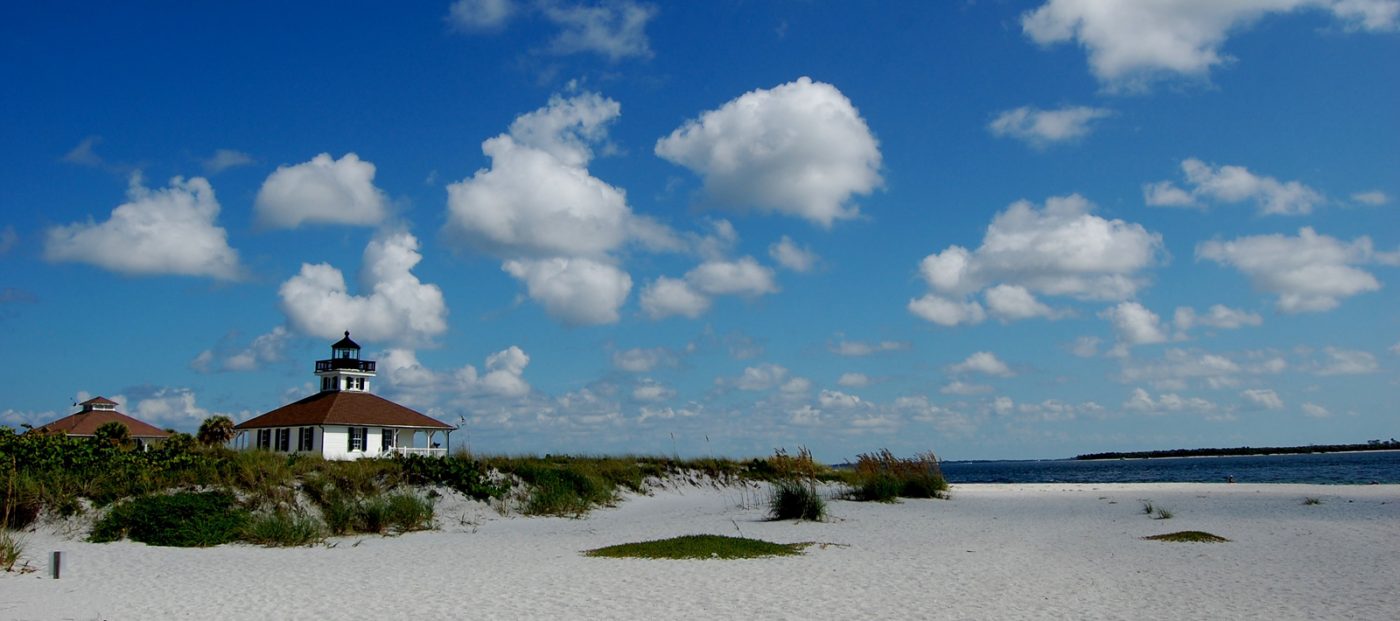 Wide-angle view of white beach, with grasses and a two-story lighthouse in the background